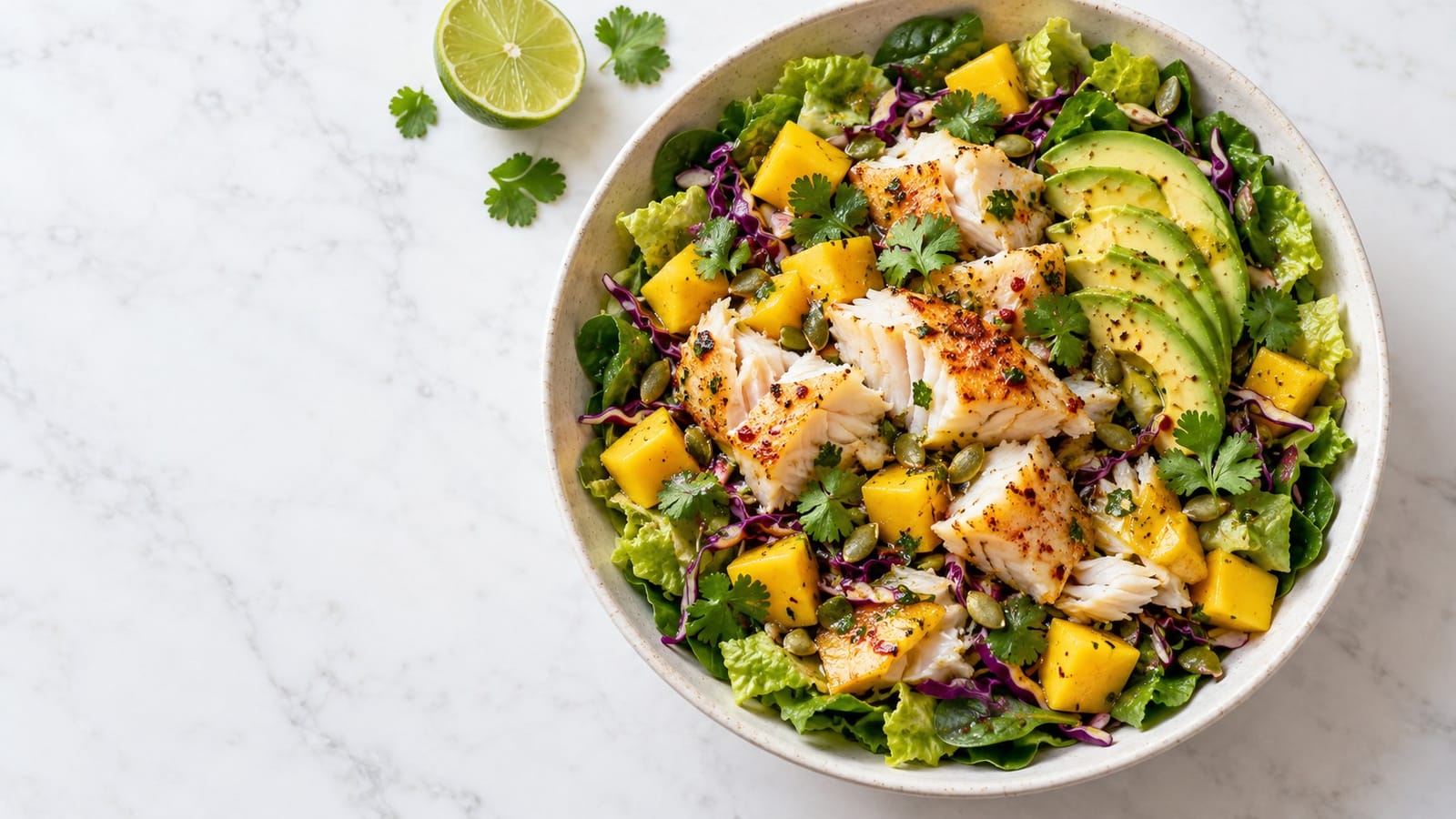 Flaked chili-lime cod with mango, avocado, romaine, spinach, purple cabbage, cilantro and pumpkin seeds in a pale ceramic bowl.